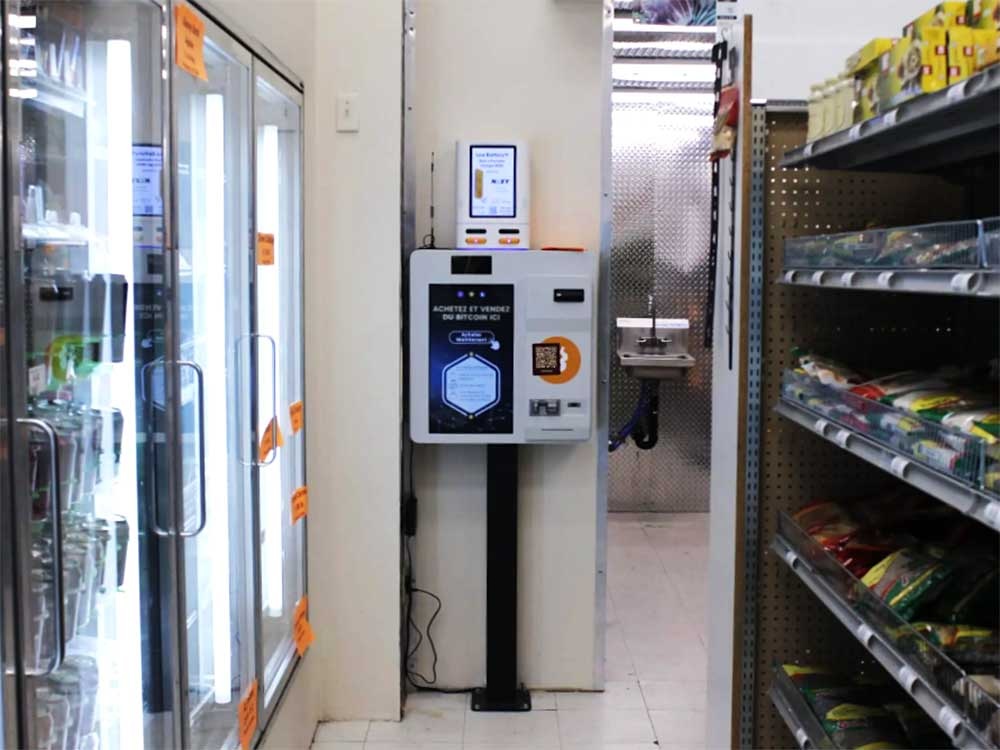 A light grey and black ATM mounted on a black pole against the wall of a convenience store, with a shelf of foodstuffs on the right and refrigerators on the left.