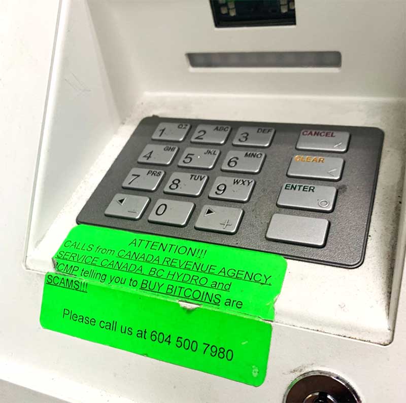 A green sign under an ATM keypad reads 'Attention!!! Calls from Canada Revenue Agency, Service Canada, BC Hydro and RCMP telling you to buy bitcoins are scams!!!'