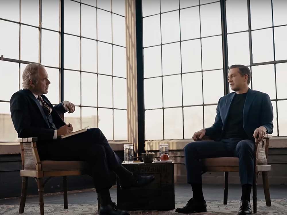 A 62-year-old light-skinned man wearing a suit sits in one chair, facing a 45-year-old light-skinned man wearing a black T-shirt and blue blazer. They are in front of a wall of windows.