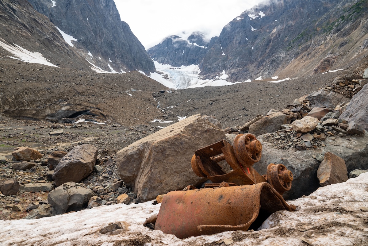 In the foreground, old, rusted-out mining equipment is clustered around a large rock. In the background, a receding glacier takes up the space between two mountain peaks.