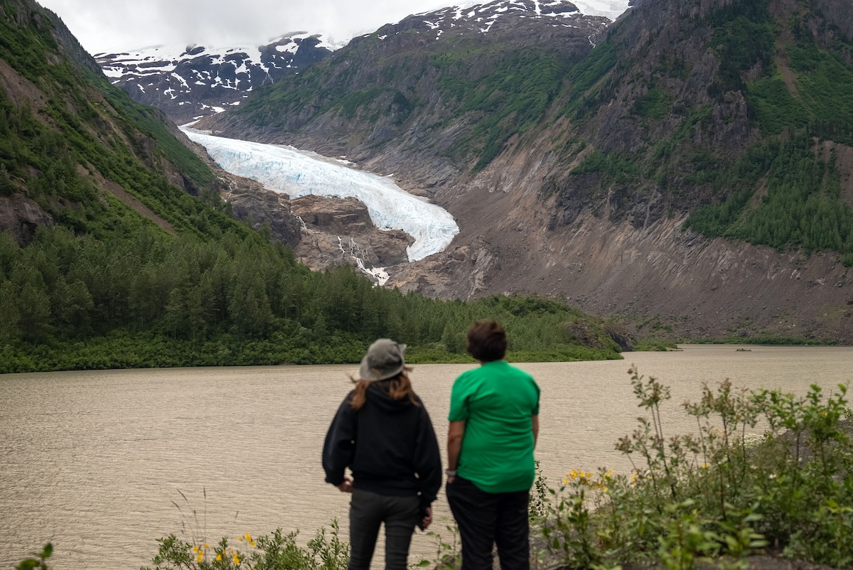 From the rear: two people look across a wide, muddy river towards a glacier.