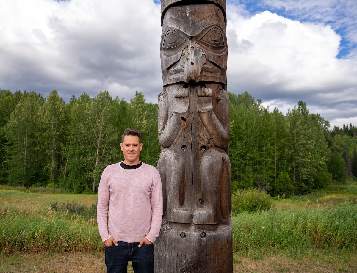 A man wearing a pink shirt and blue jeans stands next to a totem pole. Behind him, tall grass and a forested area.
