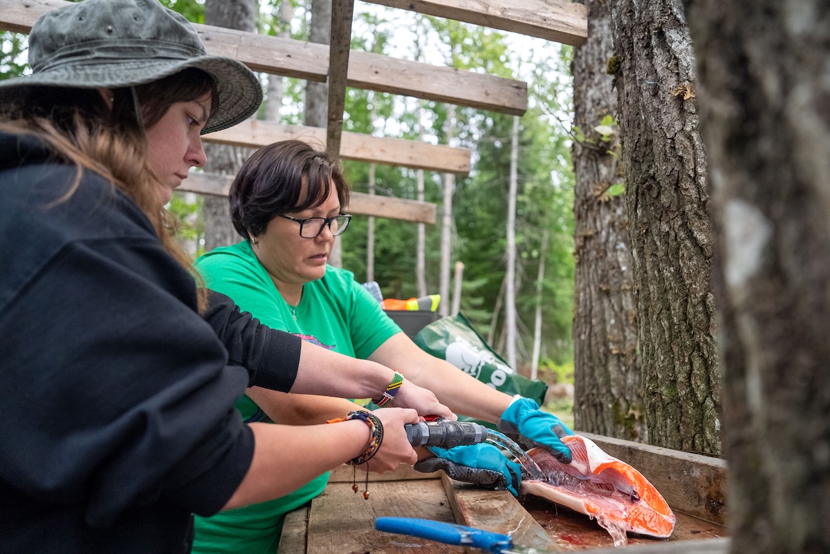 Two women stand by a wooden table. One holds open a salmon that has been deboned while the other sprays water into the open salmon.