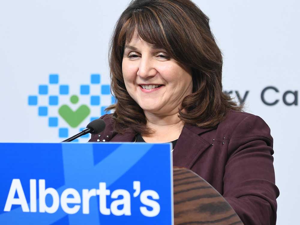 A 64-year-old light-skinned woman with long brown hair smiles as she stands at a podium.