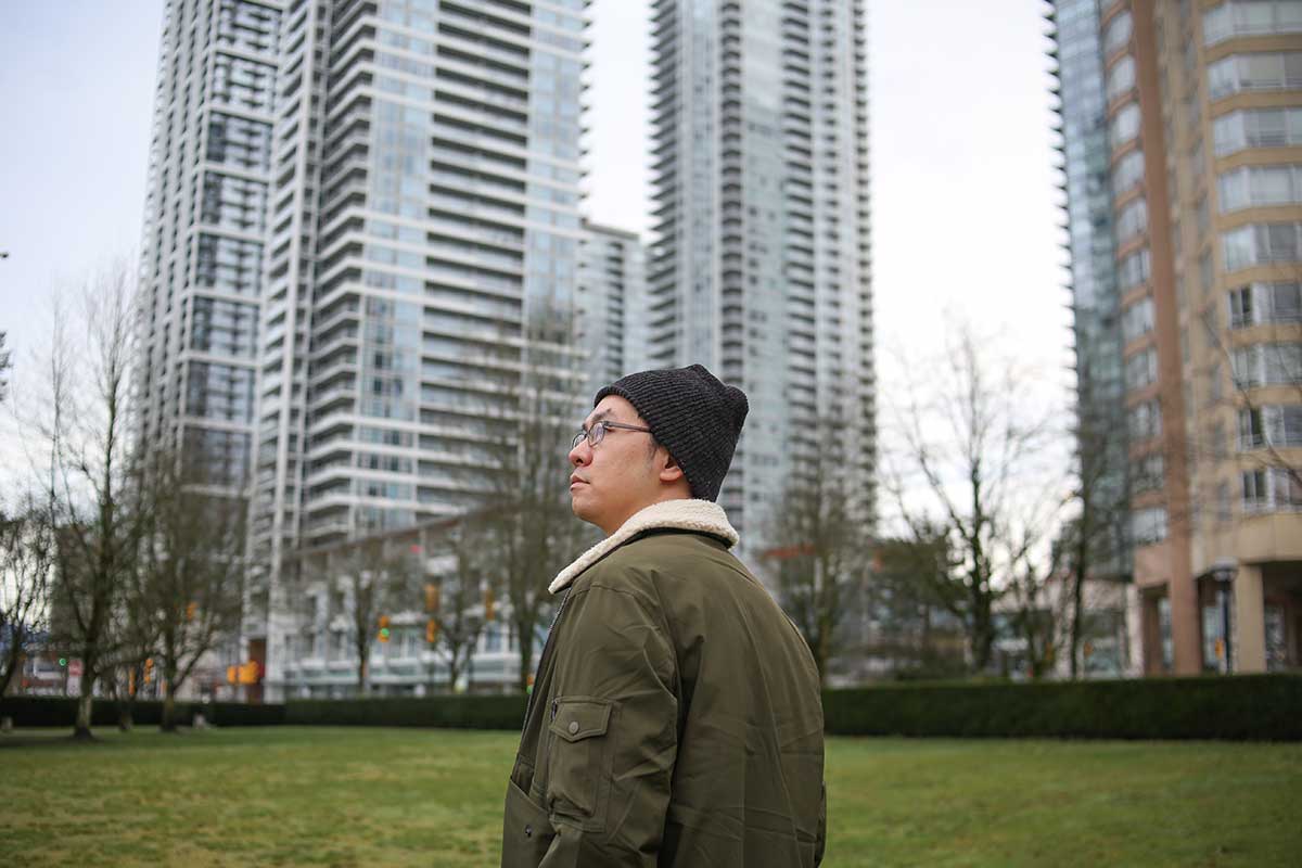 Howard Chai is standing in a green space surrounded by highrise towers on a winter day. He is turning towards the left of the frame and is seen in profile. He wears a black toque, glasses and an olive jacket with a shearling collar.