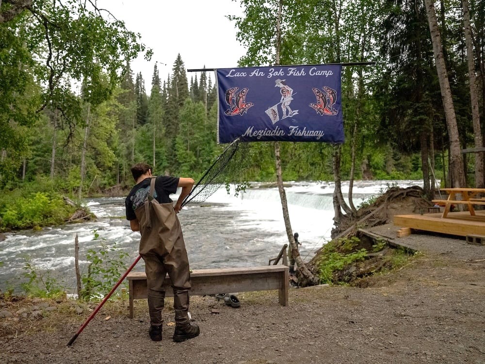 A man wearing waders puts a dip net together by the river. Behind him, a flag reads, 'Lax An Zok Fish Camp. Meziadin Fishway.'