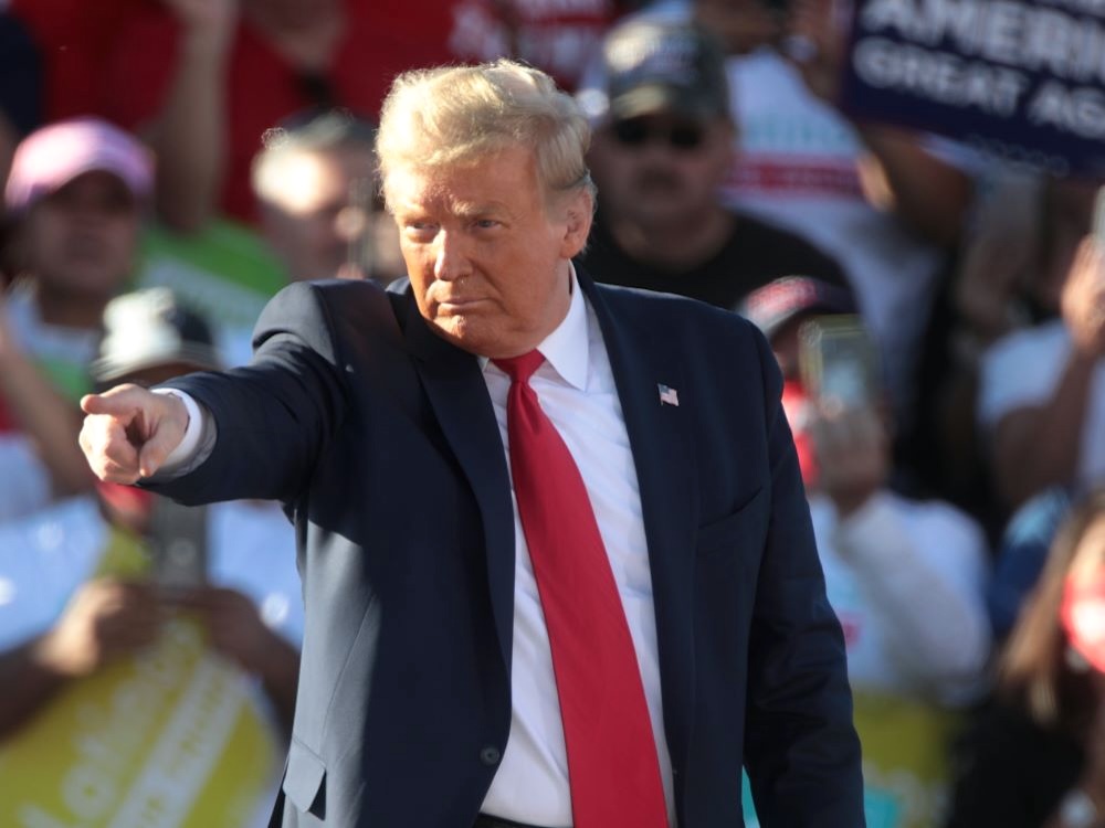 Donald Trump, a heavyset man with a reddish blond comb-over wearing a blue suit and tie, points from a rally stage.