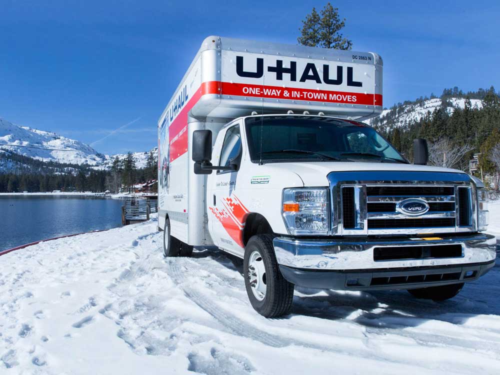 A large moving truck with the 'U-Haul' company logo is parked next to a lake in a snowy landscape with mountains in the background.