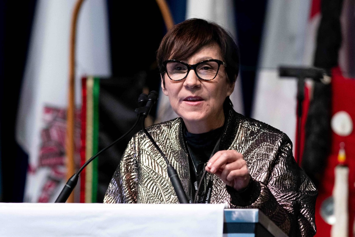 Cindy Blackstock has short dark hair and glasses. She is wearing a gold jacket with a leaf pattern over a black top. She is at a podium speaking into two black microphones. A row of flags stands behind her.