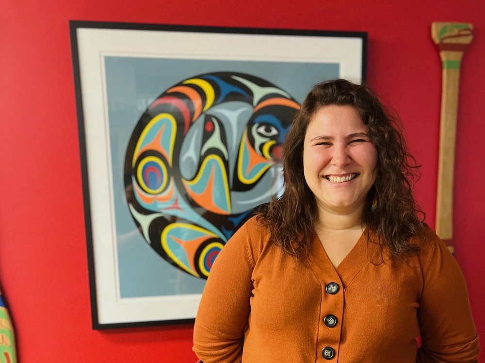 Jessica Knutson is smiling widely. She has brown, curly, shoulder-length hair and is wearing a pumpkin-coloured top. She is standing in front of a wall painted red adorned with two pieces of Indigenous artwork.