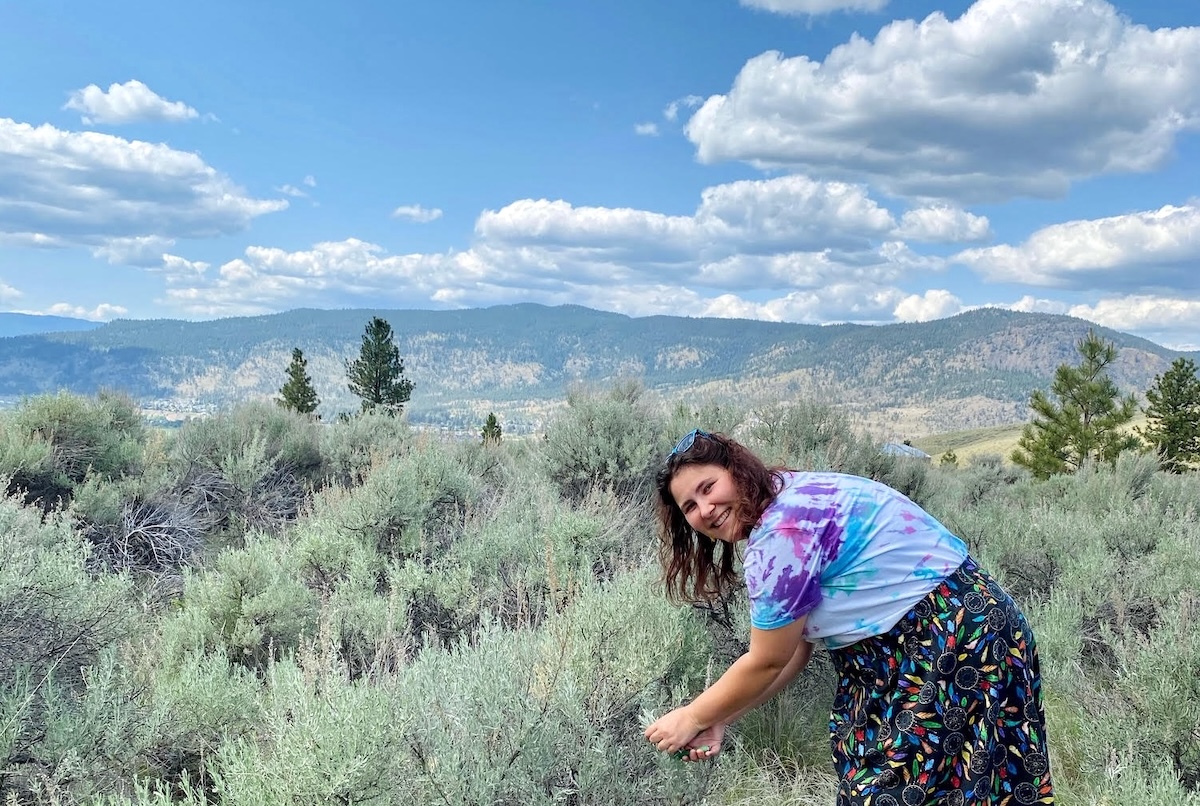 Jessica Knutson is wearing a tie-dyed T-shirt in blue, purple and white and a long black ribbon skirt featuring a dream-catcher pattern on a black background. She is picking leaves from short green shrubs in a sunny setting with a mountain in the background. She is leaning forward towards the plants and smiling at the camera.