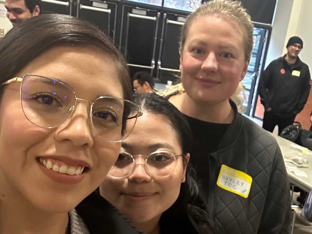 Three people look at the camera. The two on the left have medium-light skin tone and dark hair and are wearing glasses. The person on the right has light-toned skin and blond hair.