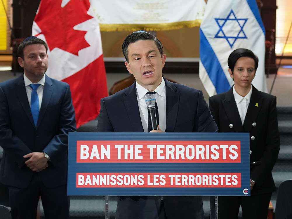 A light-skinned 45-year-old man wearing a suit and yarmulke stands at a podium that says 'Ban the Terrorists' in English and French. Behind him are Canadian and Israeli flags.