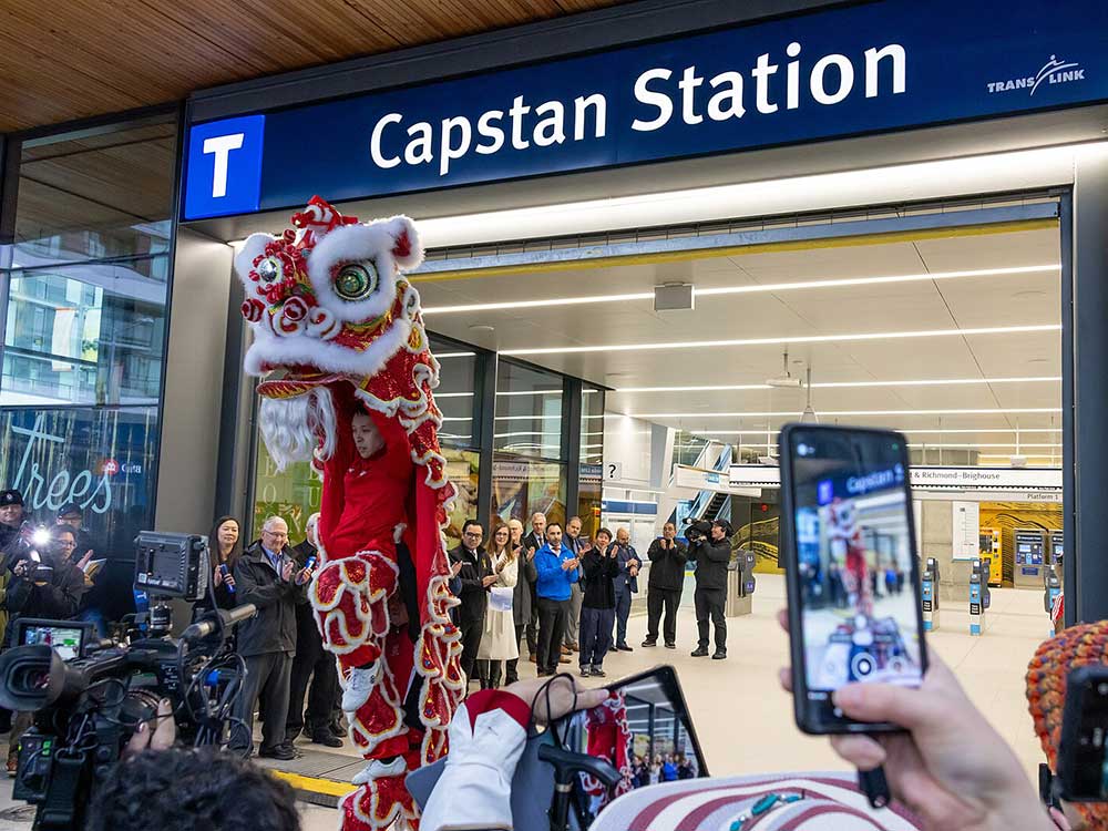 The entrance to Capstan Station bears the station’s name in sans-serif white typeface against a dark blue background with a 'T' to the left of the sign. A person holds up a red Chinese lion as part of an opening ceremony. People are applauding in the corridor behind the lion.