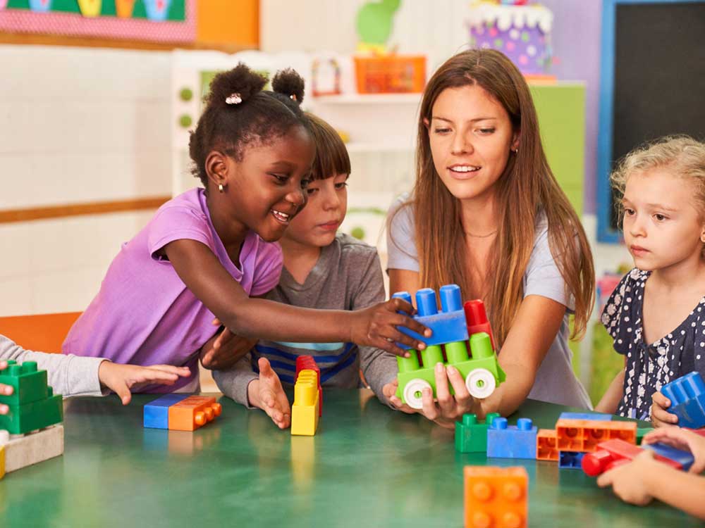 A woman with light skin tone and long brown hair plays with Duplos at a round table with a group of kids.
