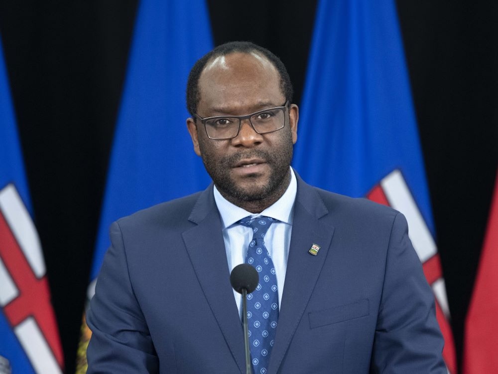 A photo of Kaycee Madu, a man with medium-dark skin tone, short hair, a short beard and glasses. Madu wears a blue suit, a blue shirt and a blue tie, and stands at a podium.