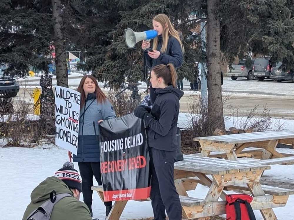 Two light-skinned women stand by a picnic table in the snow holding signs that read 'Wildfire to Wild Lies. 6 months homeless' and 'Housing Delayed = Recovery Denied.' A third woman stands on a table with a megaphone.