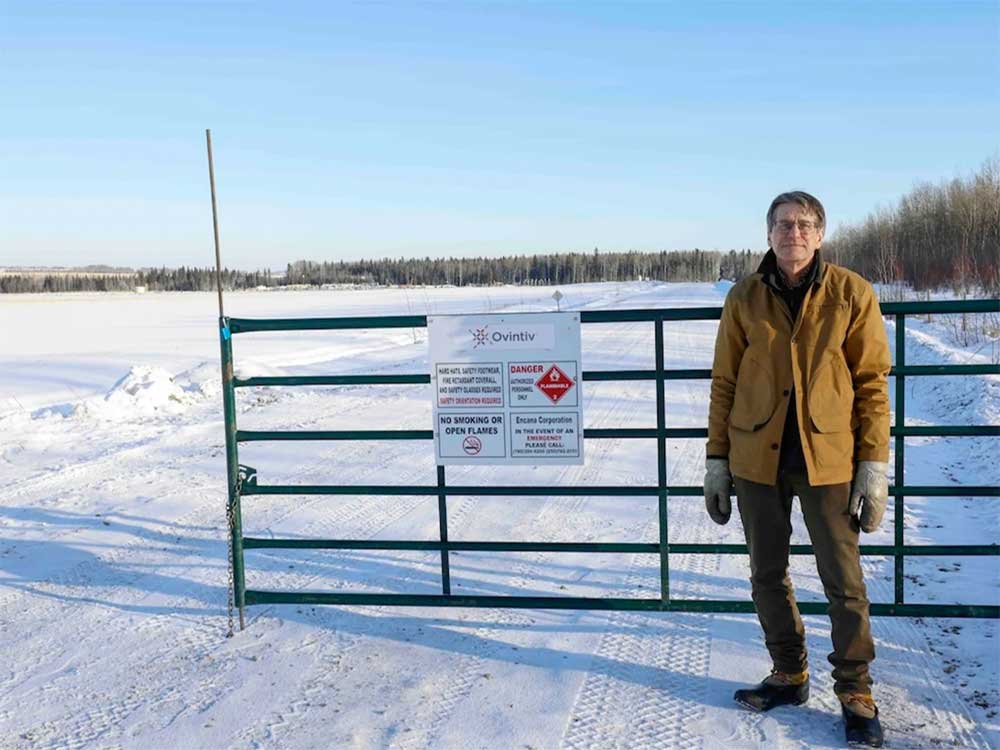 A light-skinned man in winter clothes stands beside a gate on a snowy road.