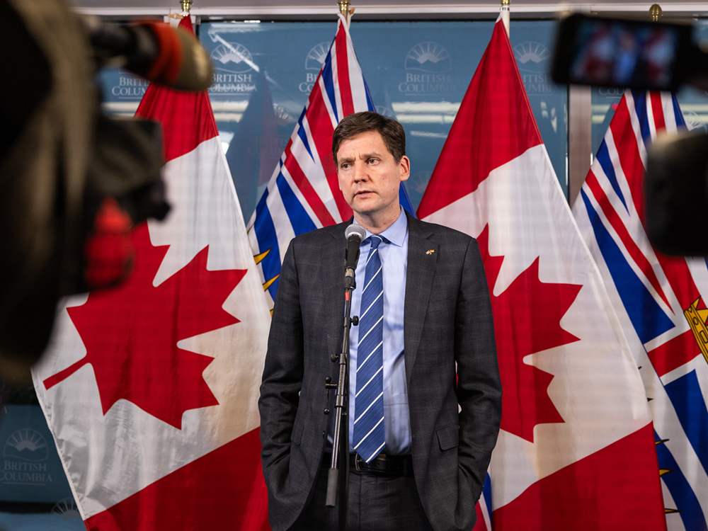 A man wearing a dark suit and tie speaks to cameras in a room with Canadian and British Columbia flags behind him.