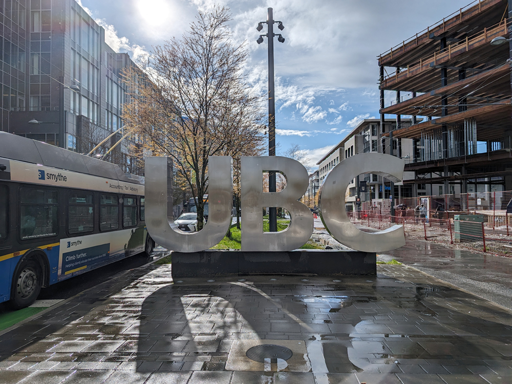 The 'UBC' sign, at the edge of campus, by the bus loop. It has just freshly rained, and 'UBC' is reflected on the shiny ground.