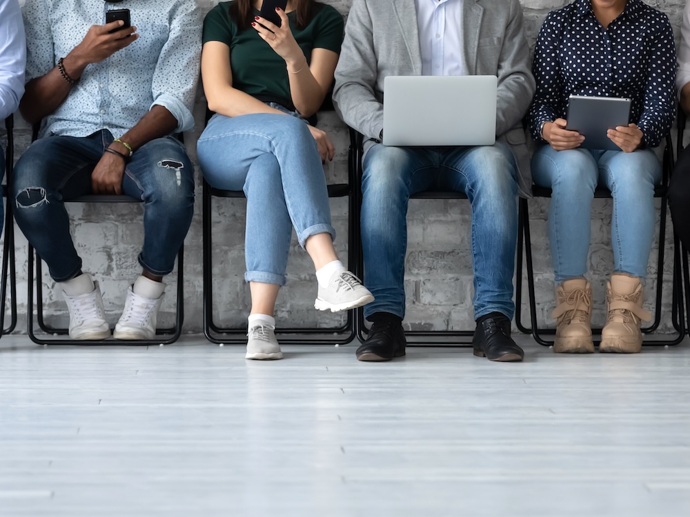 A row of youngish-looking people sit on chairs, working with tablets and laptops. Their faces aren’t visible.