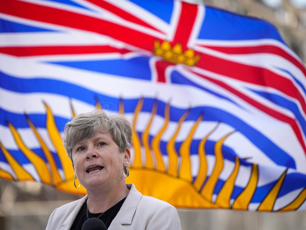 Josie Osborne, a middle-aged woman with short grey-blond hair and light skin tone, wears a black shirt and a cream-coloured blazer and gives an address in front of a British Columbia flag.