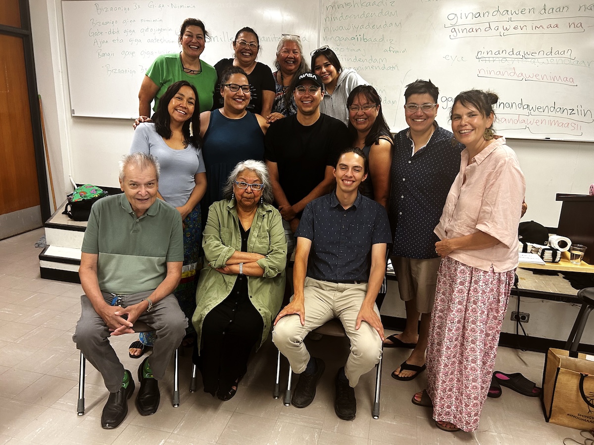 Thirteen people pose for a photo in a classroom. They are all smiling, and a range of different ages. Behind them, there are Ojibwe phrases written on the whiteboard.