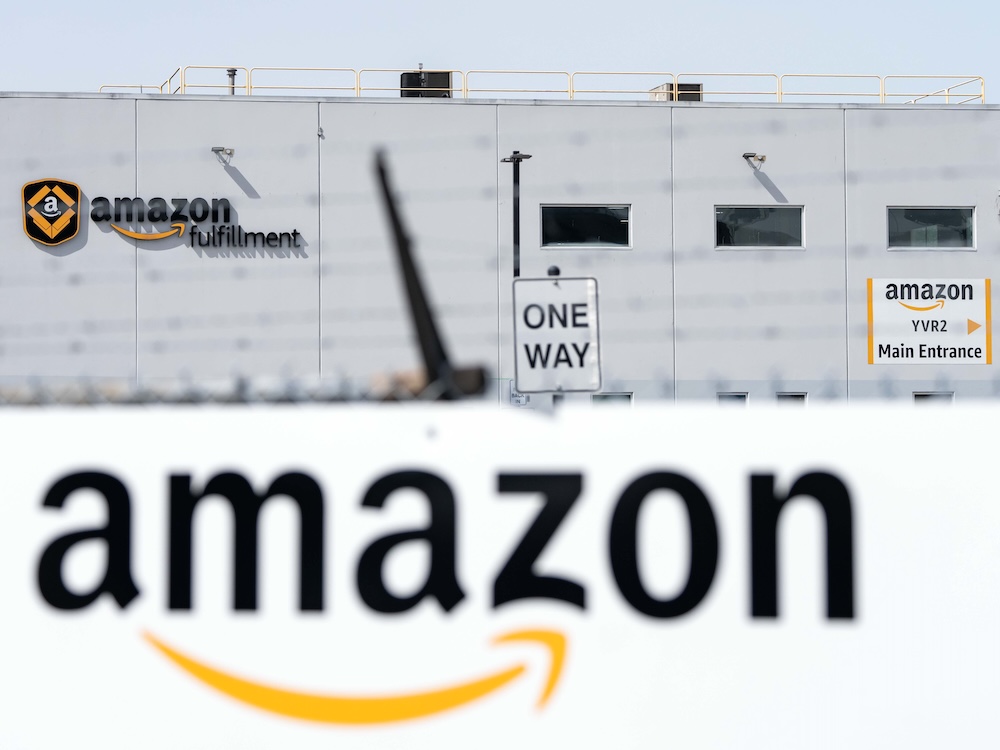 In the foreground is a sign with the Amazon logo, In the distance, a large white warehouse with another sign saying “Amazon Fulfillment.”