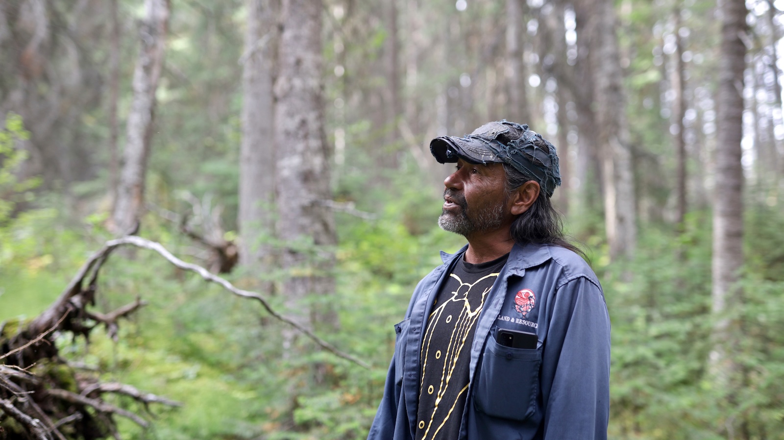 A man with medium skin tone and long dark hair wears a beat-up blue ball cap, a tuxedo T-shirt and an open blue-collared shirt with the Saik&rsquo;uz logo and 'Land & Resources.' He is standing in a forest.