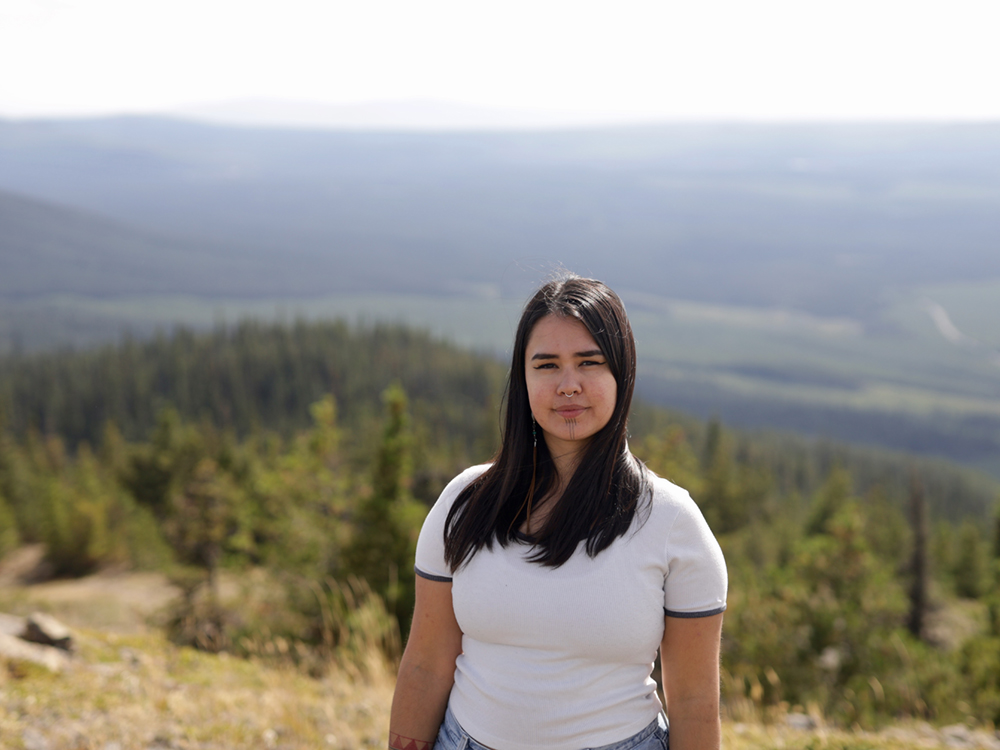 A young woman with chin tattoos, medium-light skin tone and long, straight, dark hair stands on a small mountain with a forested, logged landscape in the background.