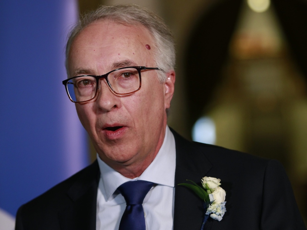 A 61-year-old light-skinned man speaks. He wears glasses, a white shirt and blue tie and a dark suit with a white flower on a lapel.