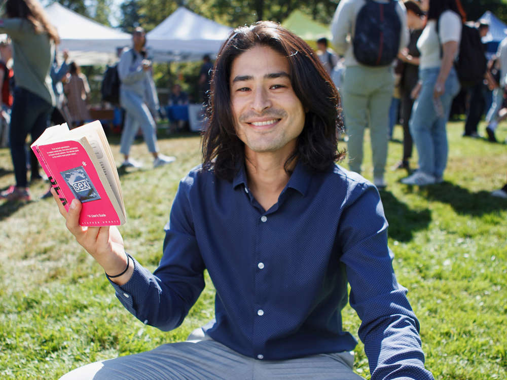 A man with thick black shoulder-length hair, a blue-collared shirt and grey pants smiles at the camera. He&rsquo;s holding a pink book and sitting on the grass. People wander across the grass in the background.