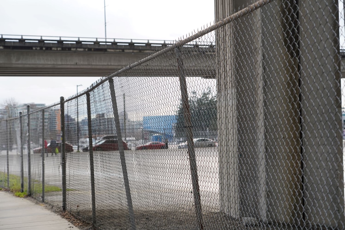 An empty parking lot is seen through a chain-link fence, with a concrete viaduct overhead.