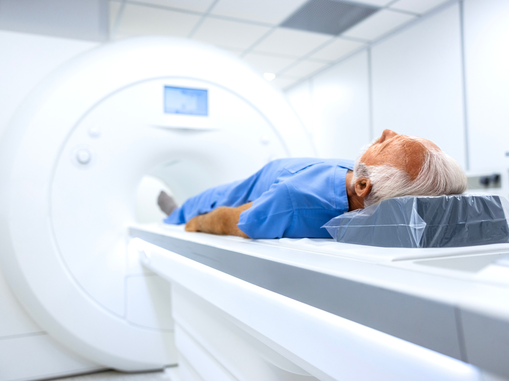 A person with a white, receding hairline gets ready for an MRI. They&rsquo;re wearing a blue medical robe and their feet are pointed towards the MRI machine.