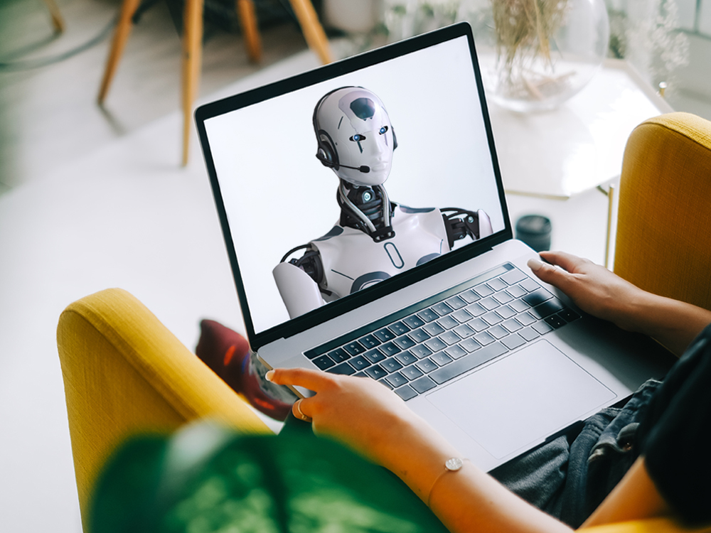 A person sits in a chair holding an open laptop. On the screen is a robot wearing a headset.