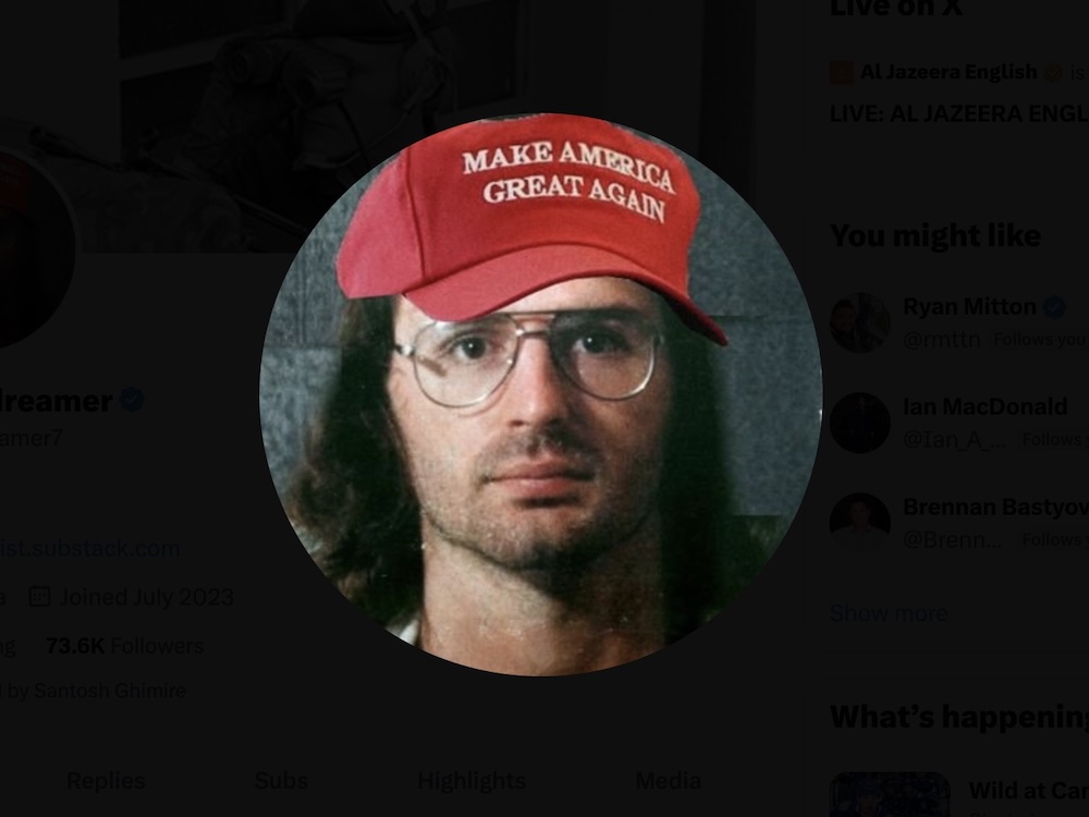 A 30-ish man with shoulder-length dark hair and stubble with a photoshopped red Make America Great Again hat.