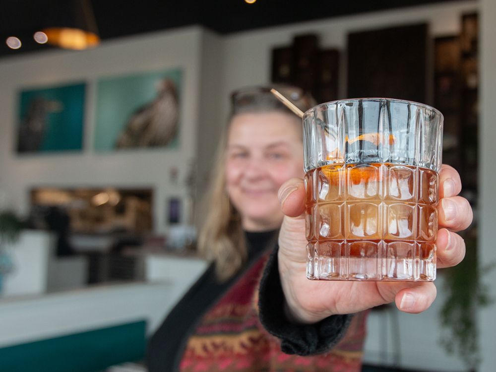 A woman with blond hair holds out a glass that holds an amber liquid with a twist of orange and cherry on a toothpick.