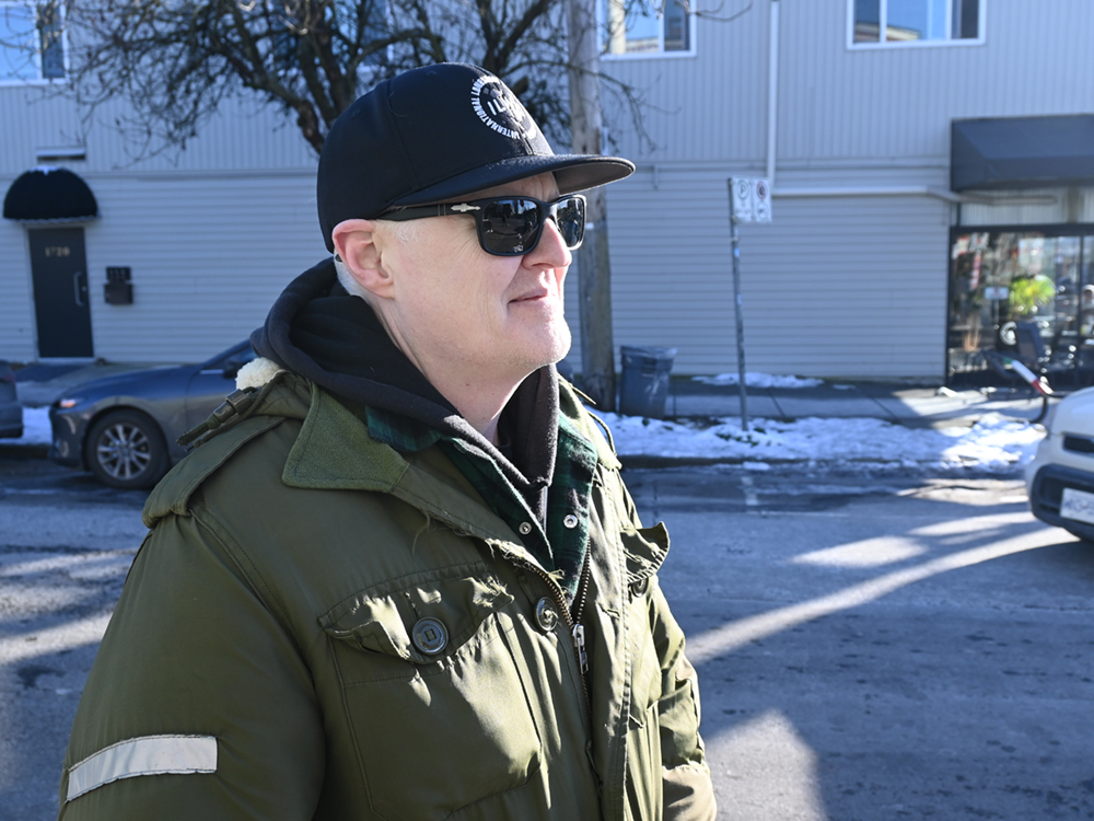 A man with pale skin wears a warm winter coat and dark sunglasses with a black hat pulled low to protect his eyes from the bright winter sun. He’s standing on a residential street looking at something to the right of the camera.