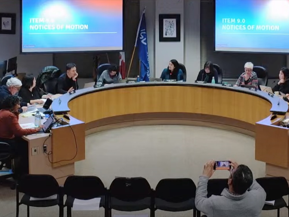 A group of school trustees sit at a semicircular meeting table. In the foreground, a man stands and records using his cellphone.