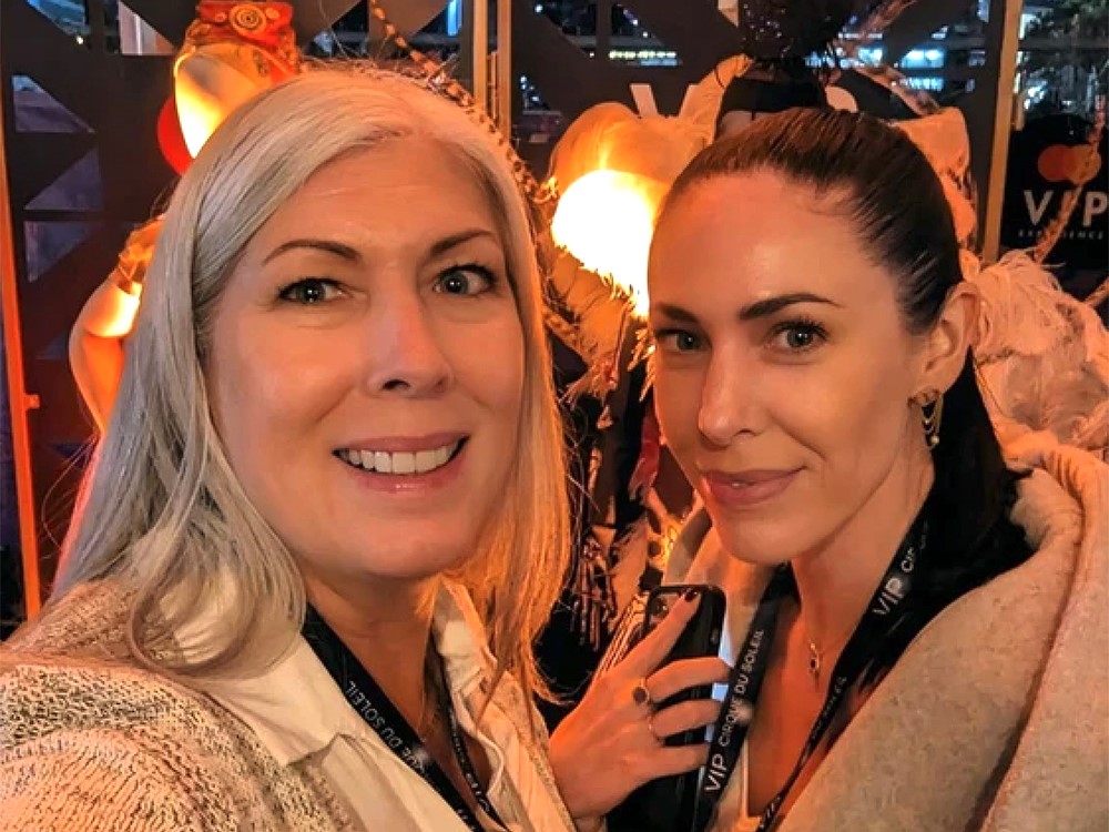 The photo is a selfie of two women. The woman on the left has silver hair and smiles at the camera. The woman on the right has dark hair pulled back into a ponytail.