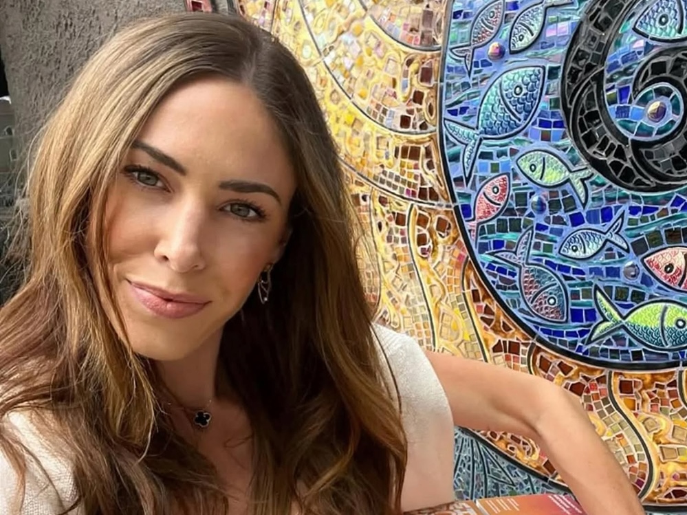 A woman with long brown hair looks at the camera. Behind her there is a colourful tile mosaic.