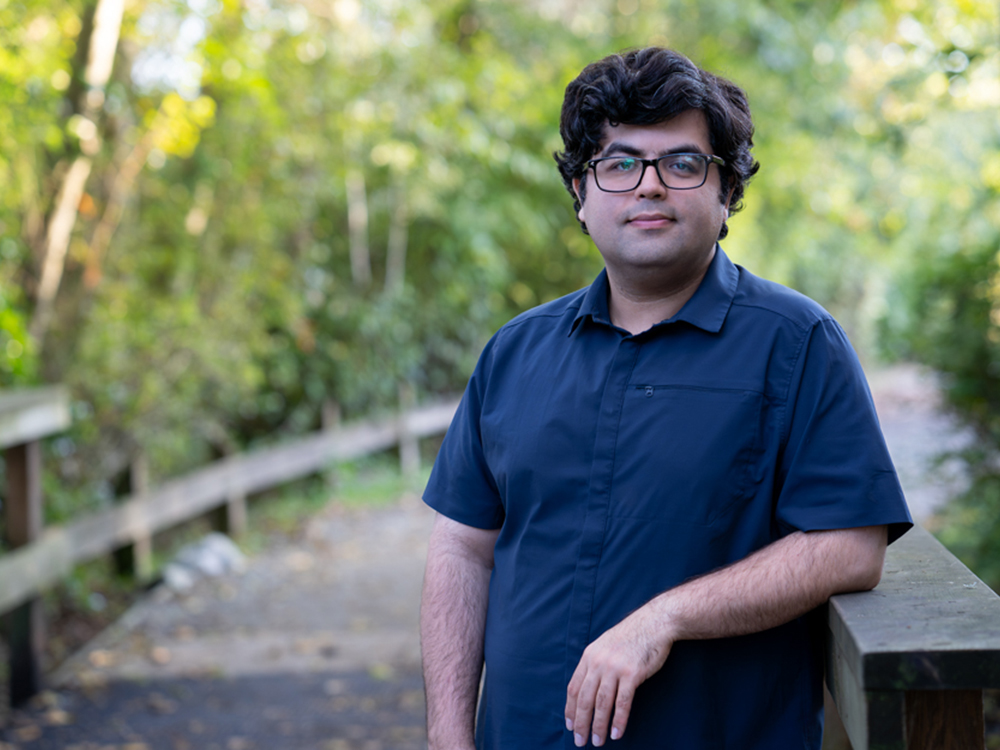 A man wearing a blue button-down T-shirt and glasses leans against a wooden handrail on a scenic path in the woods. He is clean-shaven, has medium-light skin tone and thick dark hair. His expression is relaxed and friendly.