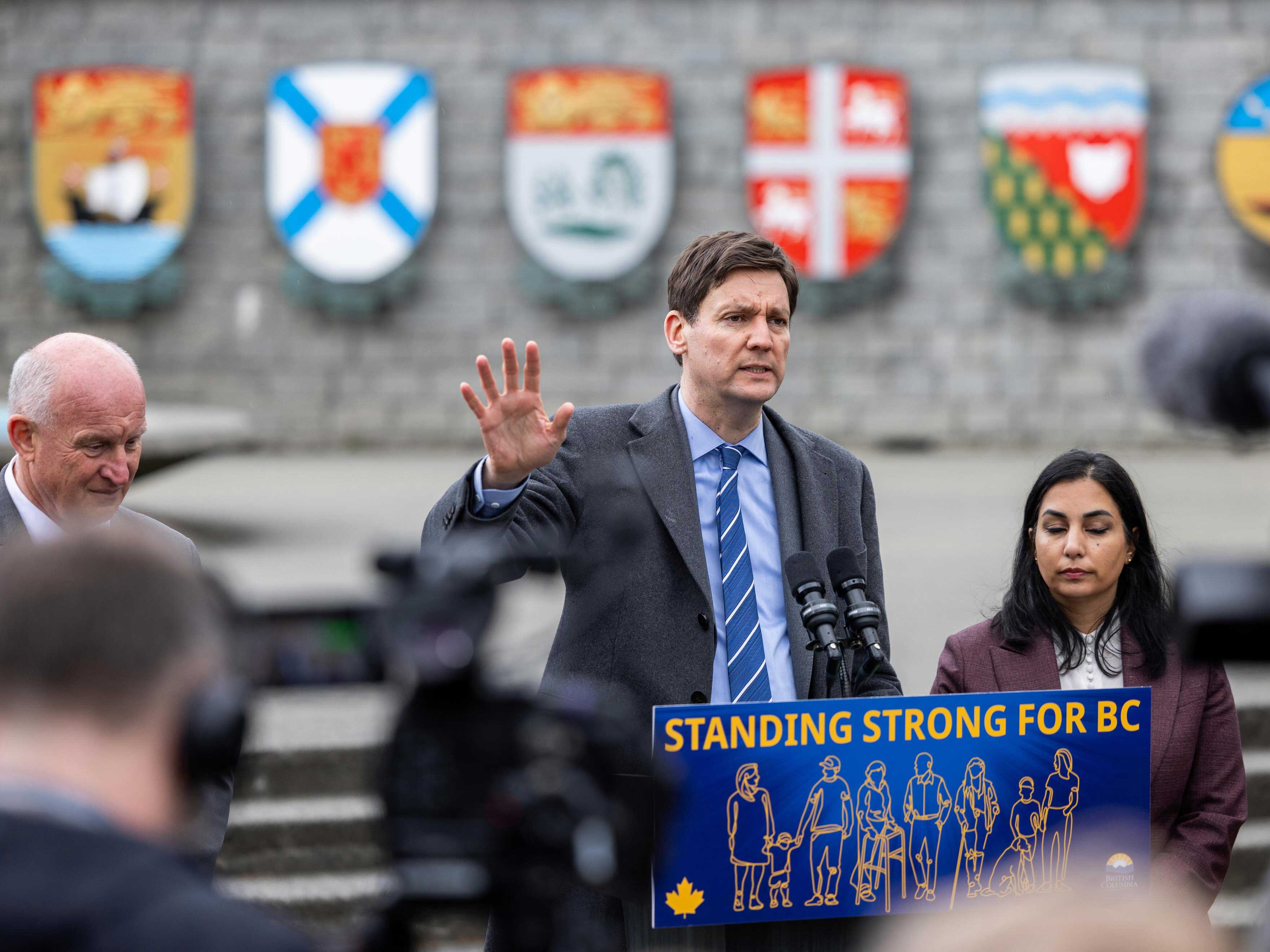 David Eby, a tall light-skinned man, stands at a microphone. Attorney General Niki Sharma stands to his left.