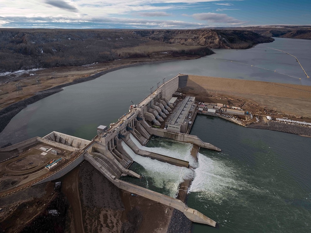 An aerial photo shows the Site C dam across the Peace River, with water flowing over the top.