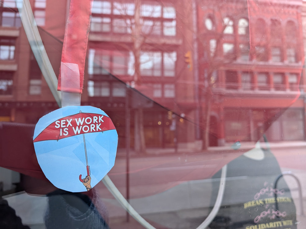 A sticker with a red umbrella reading, ‘Sex work is work’ has been stuck to a window. The window reflects DTES buildings across the street.