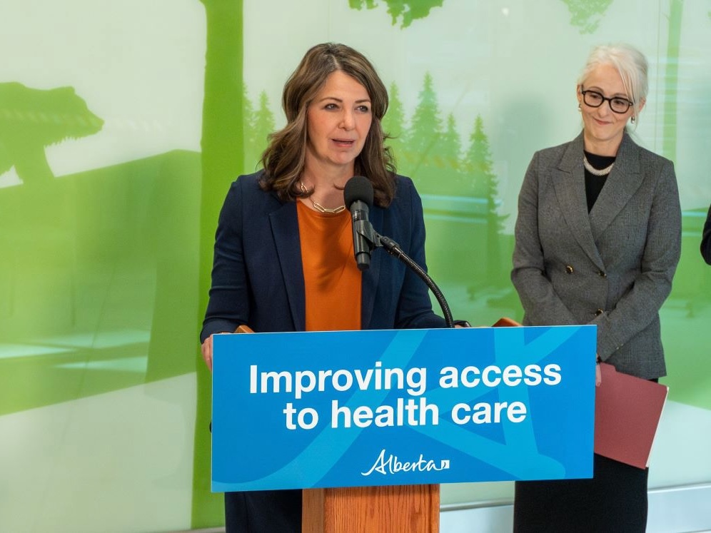 Danielle Smith, a 54-year-old light-skinned woman with shoulder-length brown hair, stands at a podium where a sign says &lsquo;Improving Access to Health Care.&rsquo; Another light-skinned woman looks on, holding a folder.