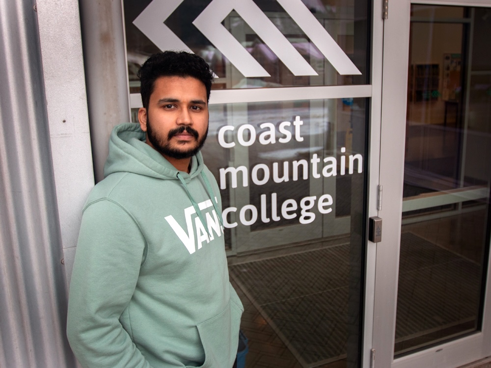A man with medium-dark skin tone, short black hair and a beard, and wearing a hoodie, stands in front of glass doors with a logo that says &lsquo;Coast Mountain College.&rsquo;