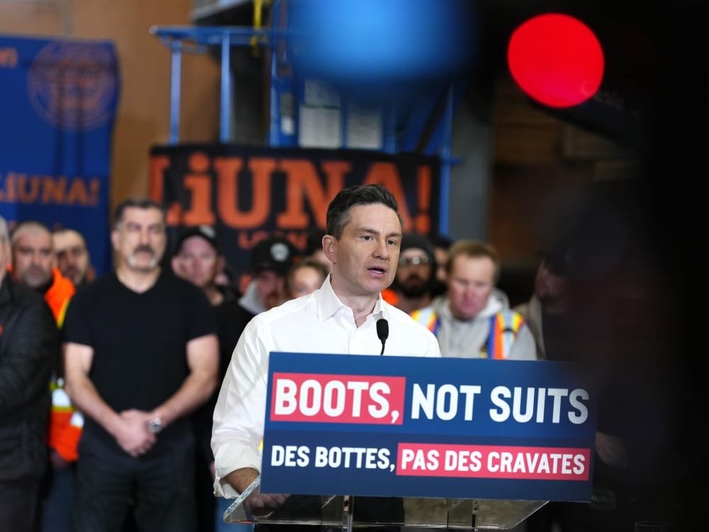 Pierre Poilievre, a 45-year-old light-skinned man, stands at a podium with people behind him. A sign says, ‘Boots, Not Suits.’