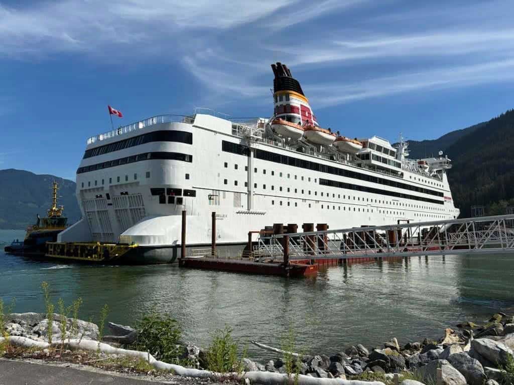A large white converted cruise ship is docked, with green hills across the water.