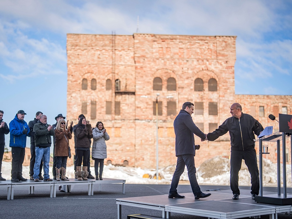 Two men shake hands while standing on a riser in an open concrete area. Behind them are several people standing on a separate riser, and a brick building.